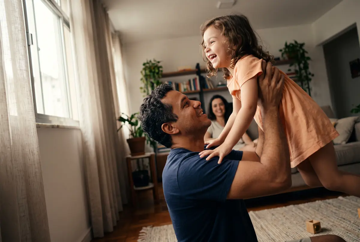 Pai brincando com a filha pequena na sala de casa, com a mãe sorrindo no sofá